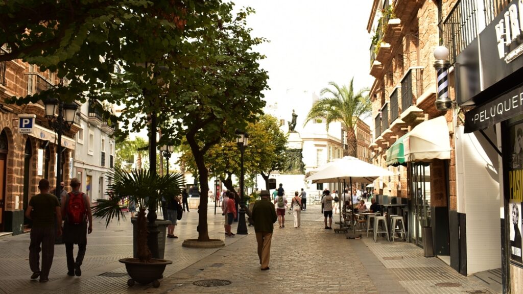 Calle arbolada en Cádiz con peatones, sombra y pavimento de piedra.